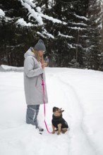 A woman in a warm coat and hat walks her dog on a snowy trail. The forest around is covered in