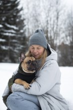 A woman in winter attire lovingly holds her French Bulldog in a snowy landscape. The scene captures