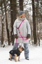 A woman in a winter coat and hat walks a French Bulldog through a snowy forest. The dog wears a