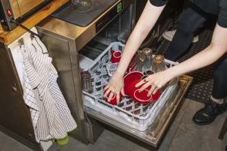 A person loads a dishwasher with red coffee cups and glass bottles, capturing the behind-the-scenes