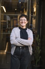 Friendly barista smiling confidently outside a cozy specialty coffee shop. Glass door entrance with