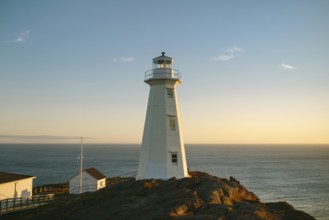 A warm sunset casts a golden glow over the white structure of Cape Spear Lighthouse, perceived as
