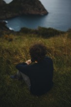 Back view of an unrecognizable man seated on grassy terrain, gazing towards a distant sea with
