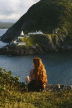 A woman with long red hair sits contemplatively overlooking the dramatic landscape of Cape Spear