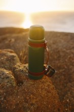 A thermos sits on a rugged rock with the warm sunset illuminating the ocean backdrop in Cape Spear