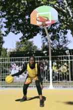 A woman athlete in a yellow jersey dribbles a basketball on an outdoor court with a colorful hoop.