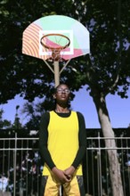 A woman basketball player in a yellow jersey stands confidently beneath a colorful hoop. The scene