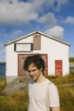 A young man with curly hair stands outdoors, smiling subtly near an old white building with red