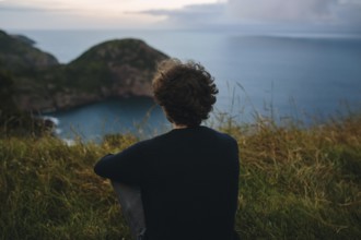 Back view of unrecognizable person sitting on grassy hill, overlooking a secluded coastal inlet