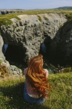 Back view of an unrecognizable redhead woman sitting on a grassy edge, overlooking a rugged coastal