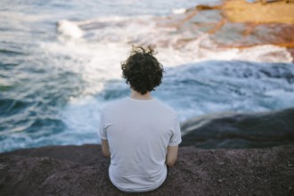 Back view of an unrecognizable person sitting on a rock by the sea, enjoying the serene view at