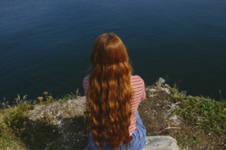 Back view of an unrecognizable redhead female sitting and looking out over the ocean from a cliff.