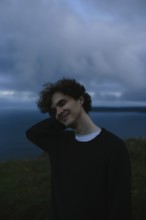 A young man stands with eyes closed relaxed against a backdrop of a cloudy sky and distant sea at