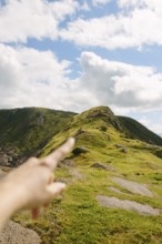 Cropped shot of unrecognizable hand pointing towards a lush green hill under a partly cloudy sky in