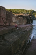 A woman in a red jacket sits pensively on a large rock cliffside, overlooking the ocean at