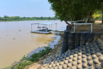 A low jetty lies on the riverbank in the shade of trees with stacked tires nearby, Porto Jofre,