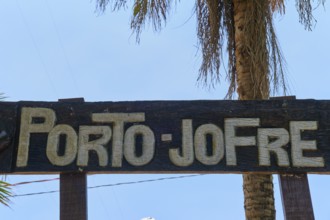 A wooden sign with the inscription 'Porto-Jofre' hangs under palm trees against a blue sky, Porto