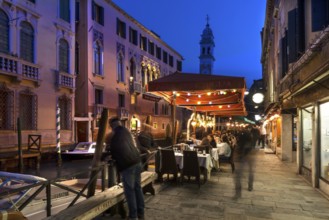 In the evening, lively restaurant on a canal with the tower of Santa Maria della Pietà in the back,