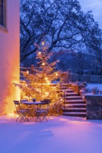 Still life with illuminated Christmas tree, stairs and table with chairs in the snow at blue hour,