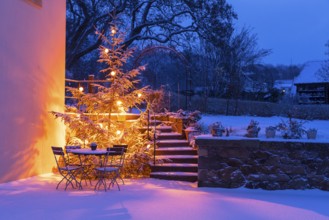 Still life with illuminated Christmas tree, stairs and table with chairs in the snow at blue hour,