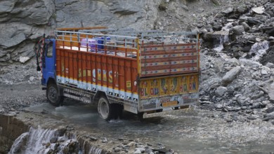 An Indian colorful truck crosses a shallow river on a road surrounded by rocky terrain on the