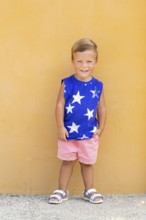 A cheerful young boy in a blue shirt with white stars poses against a vibrant yellow wall His pink