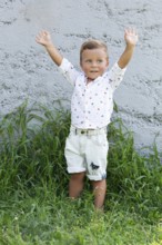 A cheerful young child stands with arms raised in a grassy garden Dressed in shorts and a patterned