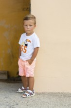 A young boy stands thoughtfully against a pastel wall, wearing a white toucan t-shirt, pastel