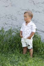 A young boy stands against a gray concrete wall, dressed in a white shirt and shorts The grass at