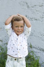A cheerful child stands against a textured wall, hands on head, wearing a white shirt with small