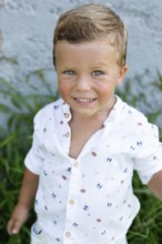 A cheerful young boy in a patterned white shirt smiles brightly at the camera while standing
