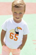 A cheerful child wearing a white T-shirt with a toucan design and number, standing outdoors on a