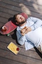 A man lies on wooden decking, enjoying a burger with a tray of fries and a drink resting on a
