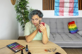 A young transgender man thoughtfully applies makeup, sitting at a table with various makeup items.