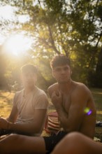 Two young men relax near a serene lake during summer, basking in the golden sunlight filtering