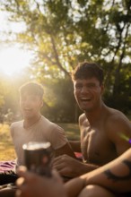 Two young men sit laughing by a lake during summer, one holding a drink. Warm sunlight illuminates