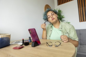 Transgender man applying makeup while recording a tutorial with a smartphone placed on a stand,