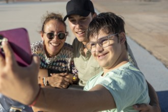 A Down syndrome teenager joyfully taking a selfie with family while enjoying a sunny day outdoors,