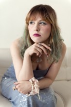 A woman with green-tinted hair and expressive makeup is seated on a white couch She wears a blue