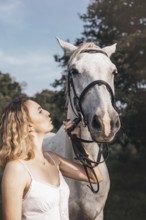 A young woman in a white dress shares a serene moment with a white horse outdoors. The sunlight