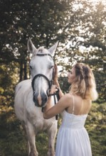 A young woman in a white dress gently caresses a white horse in a sunlit forest. The warm sunlight