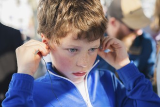 Close-up of a young boy in a blue jacket, adjusting earphones, deeply focused, sitting in a tourist