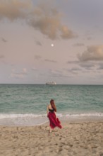 Back view of an unrecognizable redhead woman strolling along Miami Beach. She wears a flowing red