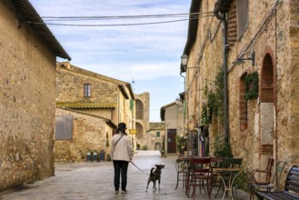 A serene street scene in Monteriggioni, Tuscany, with a woman walking her dog past rustic stone