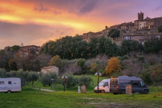 Camper vans and motorhomes parked in a scenic service area below a traditional Tuscany village. The