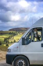 A camper van with a dog peering out the window, parked in the rolling hills of Tuscany, Italy. The