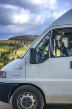 A dog looking out of a camper van window, parked amidst Tuscany's rolling hills. The landscape