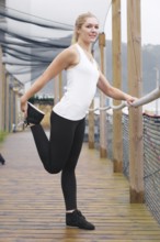 A young blond woman, dressed in athletic wear, stretches on a wooden walkway, displaying a healthy