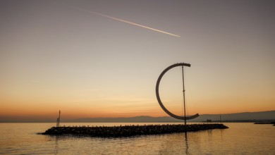 The serene scene of a sunset at Lake Geneva, showcasing a large ring-like sculpture standing on a