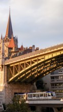 A metro train crosses a modern bridge in the foreground with the historic Lausanne Cathedral rising